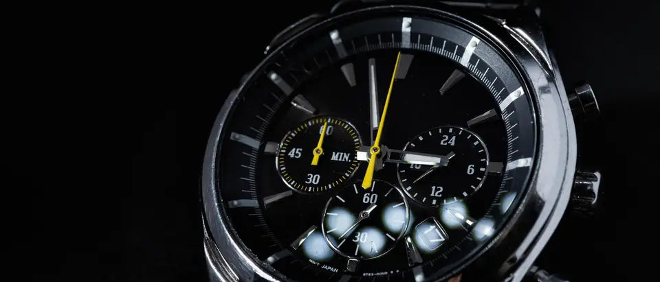 A steel chronograph wristwatch with a black dial, subdials and a yellow second hand on a black background