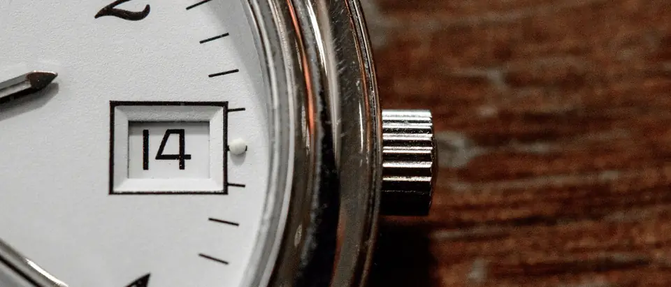 A close view of a watch with a white dial, printed numerals, a date window reading 14 and a crown, on warm wood