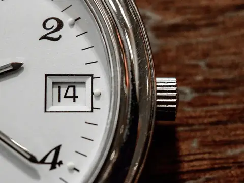 A close view of a watch with a white dial, printed numerals, a date window reading 14 and a crown, on warm wood